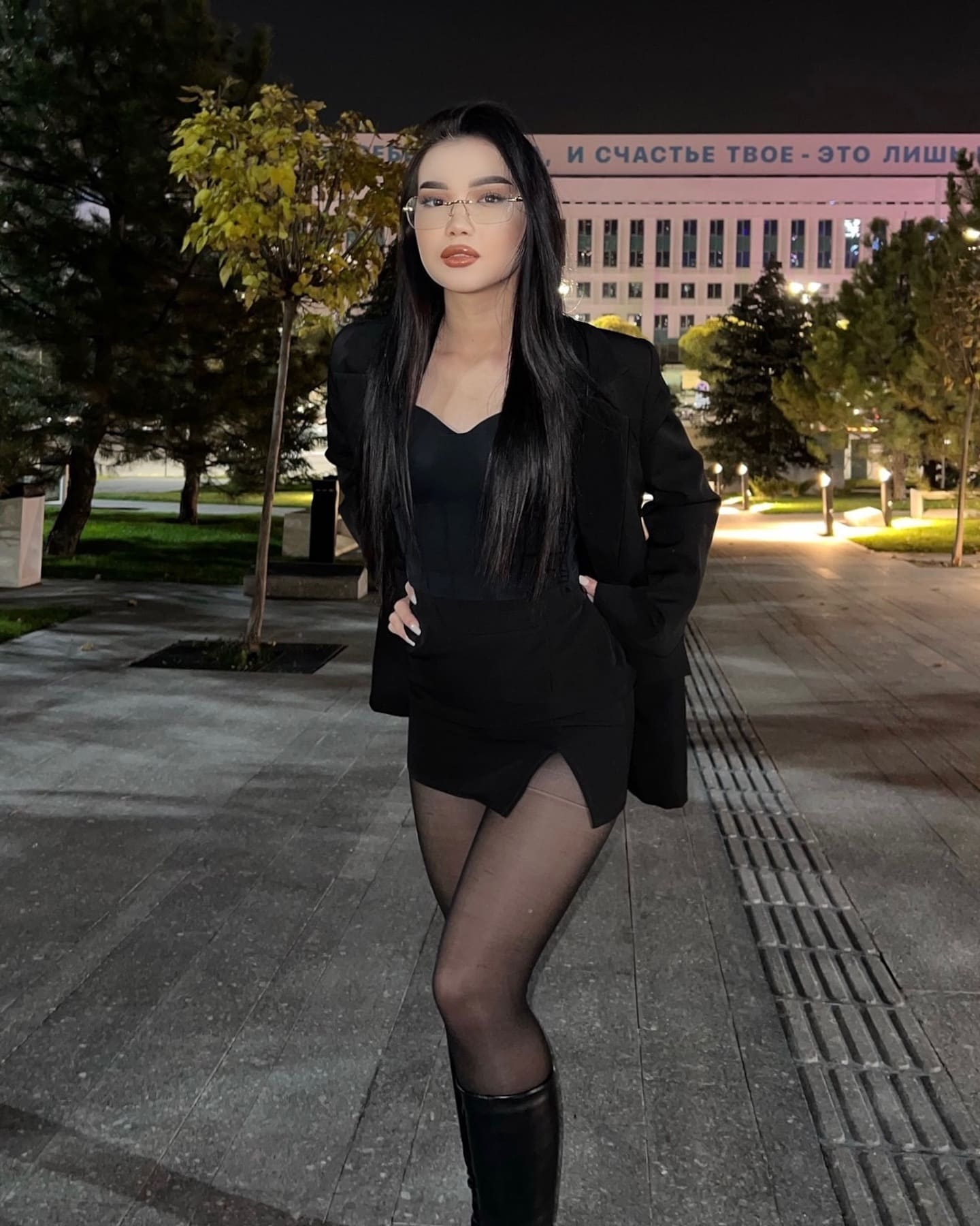 Леона в очках, черном пиджаке и платье стоит на улице ночью.